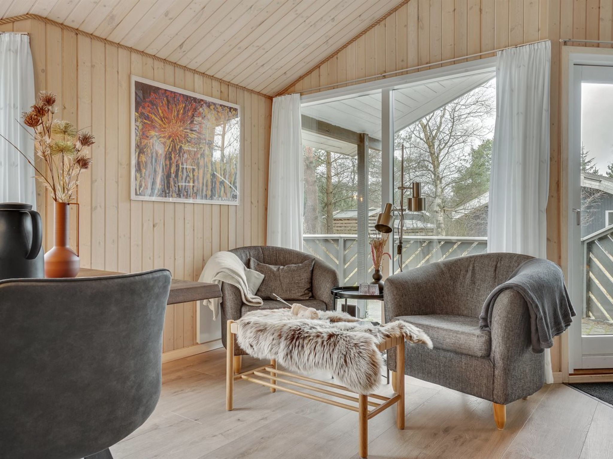 A cozy room with wooden walls, a grey sofa, a wooden coffee table with a fur throw, and a painting on the wall.