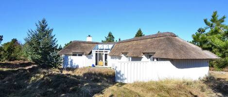 A white cabin with a thatched roof, surrounded by trees and grass.