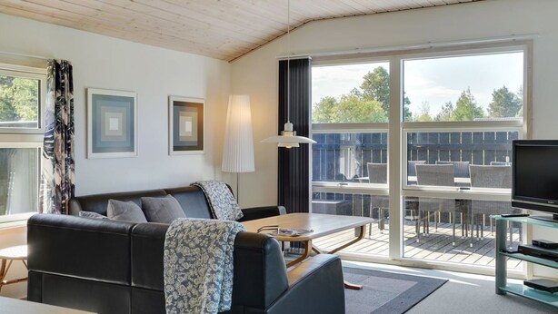 Una sala de estar moderna con un sofá de cuero negro, una mesa de centro y un mueble para la televisión. Hay ventanas amplias con vistas al exterior.