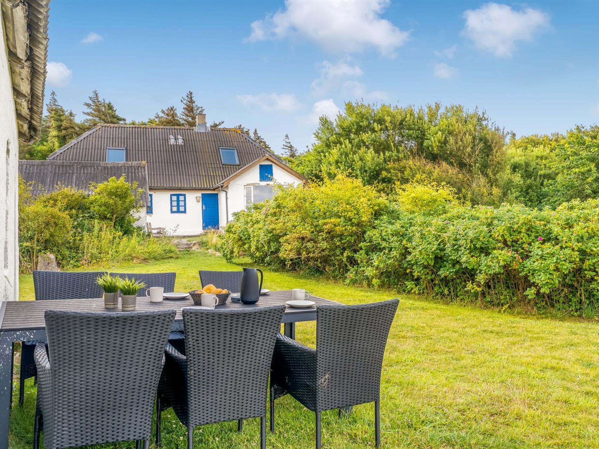 Une terrasse avec une table et des chaises, une maison avec une porte et des fenêtres bleues, et un jardin avec des arbustes et des arbres.