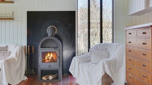 A cozy room with a wood-burning stove, two white upholstered chairs, a wooden dresser, and a window with a view of trees.