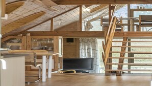 Ein geräumiges Dachzimmer mit Holzbalkendecke, einer Essecke mit Tisch und Stühlen, einem Fernseher und einem fenster mit Vorhängen.