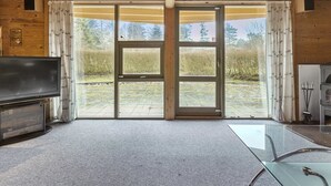 Ein Zimmer mit einer großen Schiebeglastür, die zu einem Außenbereich führt, einem Fernseher links und einem Glastisch mit Metallgestell.