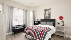 A bedroom with a bed, a bench, a nightstand, a window with curtains, and a wall-mounted artwork.