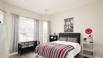 A bedroom with a bed, a bench, a nightstand, a window with curtains, and a wall-mounted artwork.