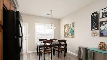 A dining area with a wooden table and chairs, a black refrigerator, a trash can, and a welcome sign on the wall.