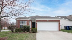 A two-story house with a brick facade, a white garage door, and a front yard with a small tree and shrubbery.