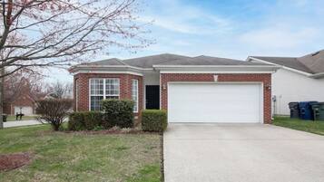 A two-story house with a brick facade, a white garage door, and a front yard with a small tree and shrubbery.