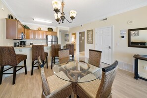 A dining area with a glass table surrounded by wicker chairs, a kitchen in the background, and a door with a decorative frame.