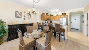 Un comedor con mesa de vidrio rodeada de sillas de mimbre, una cocina con armarios de madera y electrodomésticos de acero inoxidable, y una puerta blanca.
