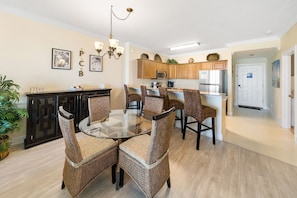 A dining area with a glass table surrounded by wicker chairs, a kitchen with wooden cabinets and stainless steel appliances, and a white door.