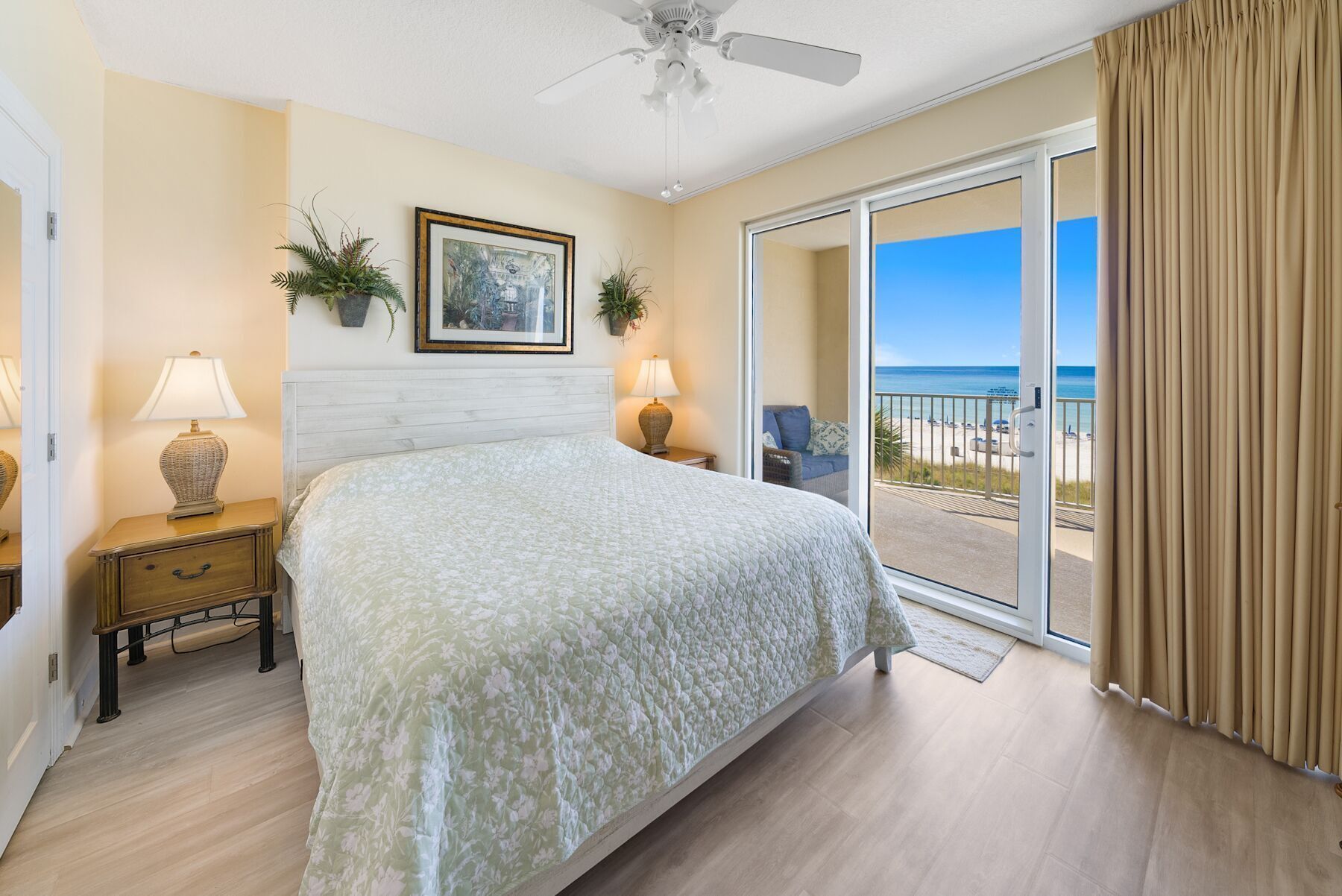 A bedroom with a bed, two bedside tables, a ceiling fan, a large window with a view of the beach, and a sliding glass door.
