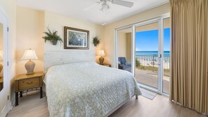 Ein Schlafzimmer mit einem Bett, zwei Nachttischen, einem Deckenventilator, einem großen Fenster mit Blick auf den Strand und einer Schiebetür aus Glas.