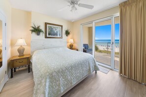 A bedroom with a bed, two bedside tables, a ceiling fan, a large window with a view of the beach, and a sliding glass door. - Salty Sundaze Beachfront King Master 2 Bed 2 Bath Sleeps 6+ Beach Service (Panama City Beach)