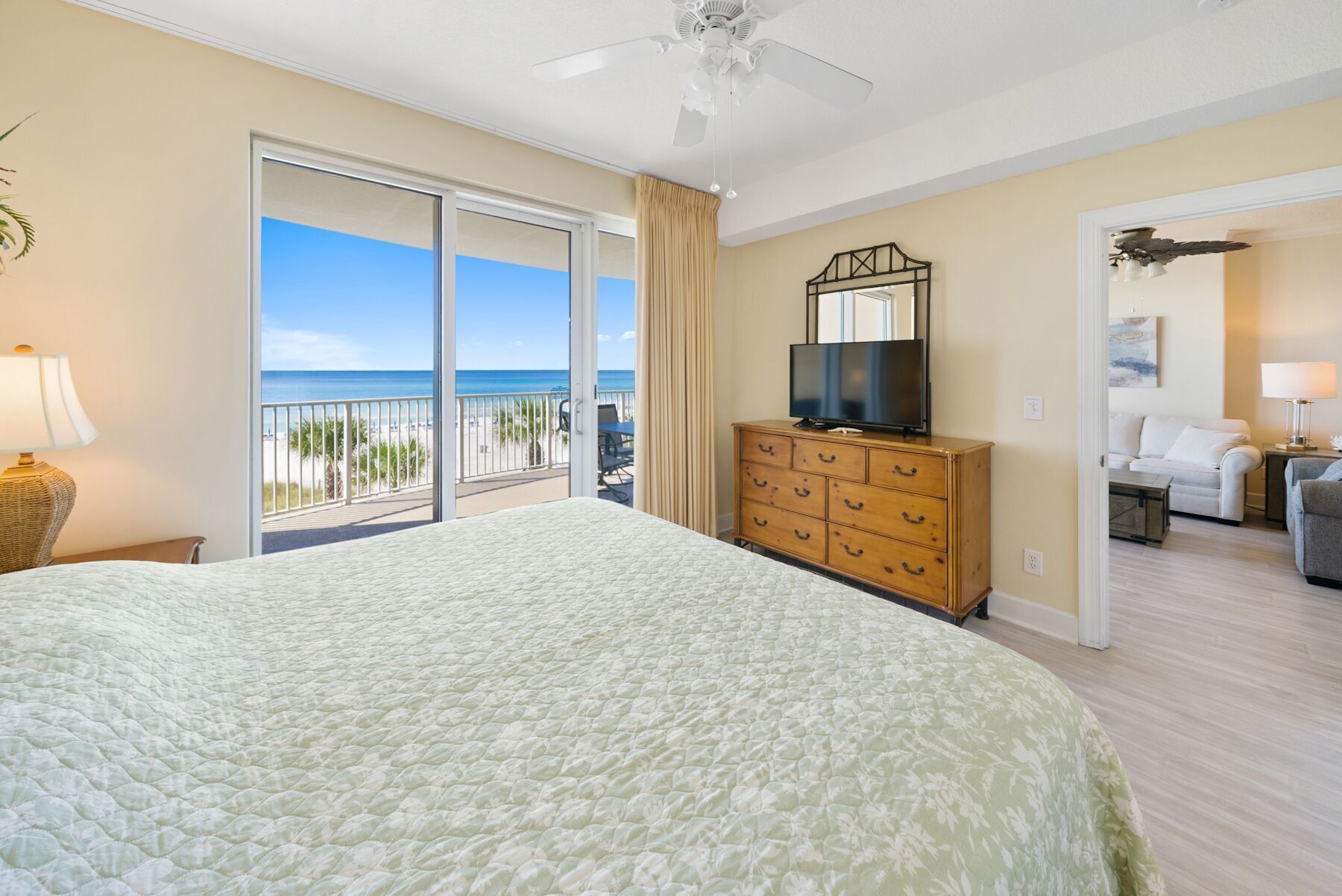 A hotel room with a large bed, a television on a wooden dresser, and a balcony with a view of the beach.