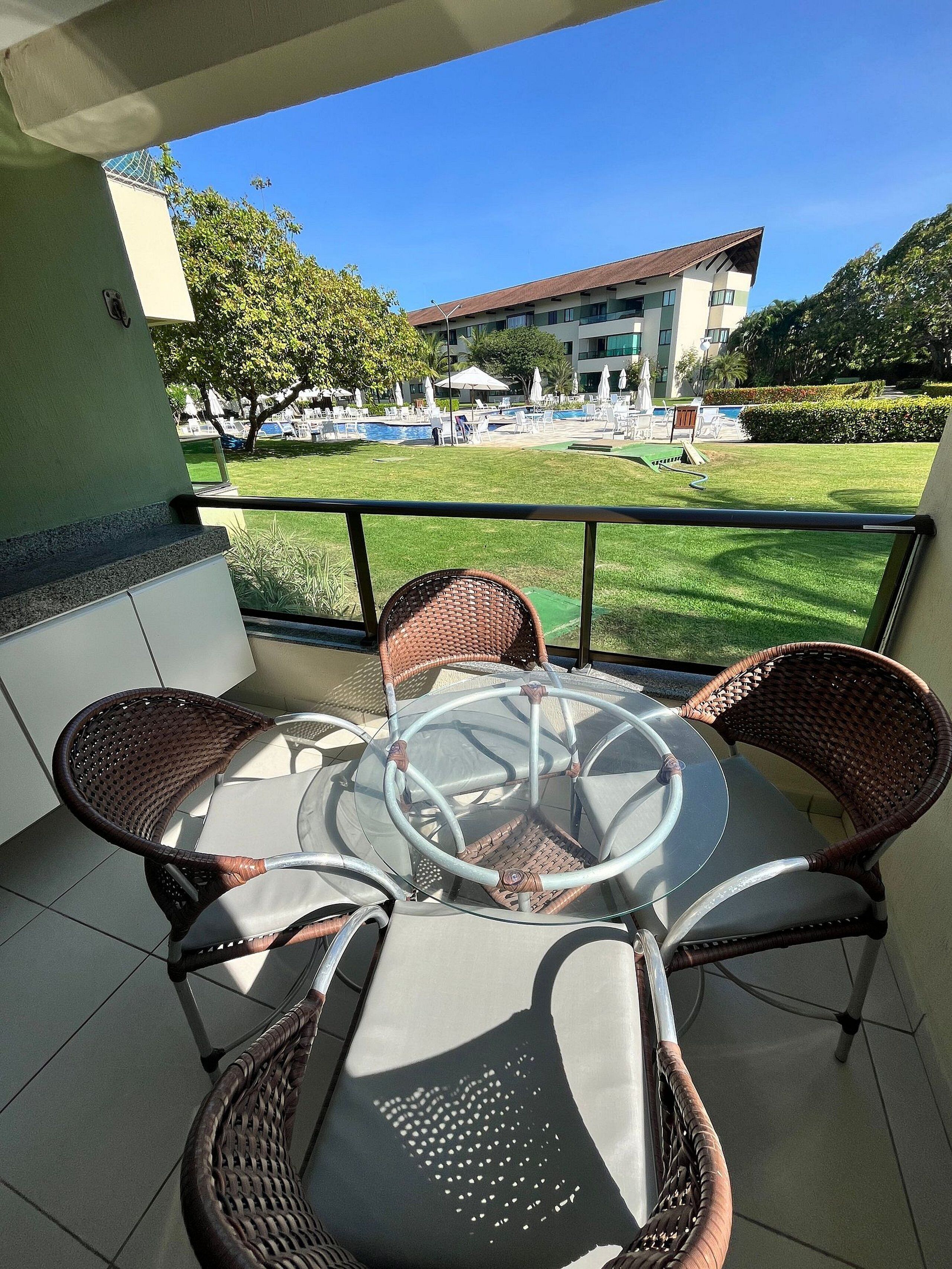 A balcony with a glass table and wicker chairs, overlooking a pool area with lounge chairs and umbrellas.