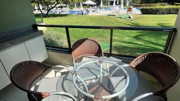A balcony with a glass table and wicker chairs, overlooking a pool area with lounge chairs and umbrellas.
