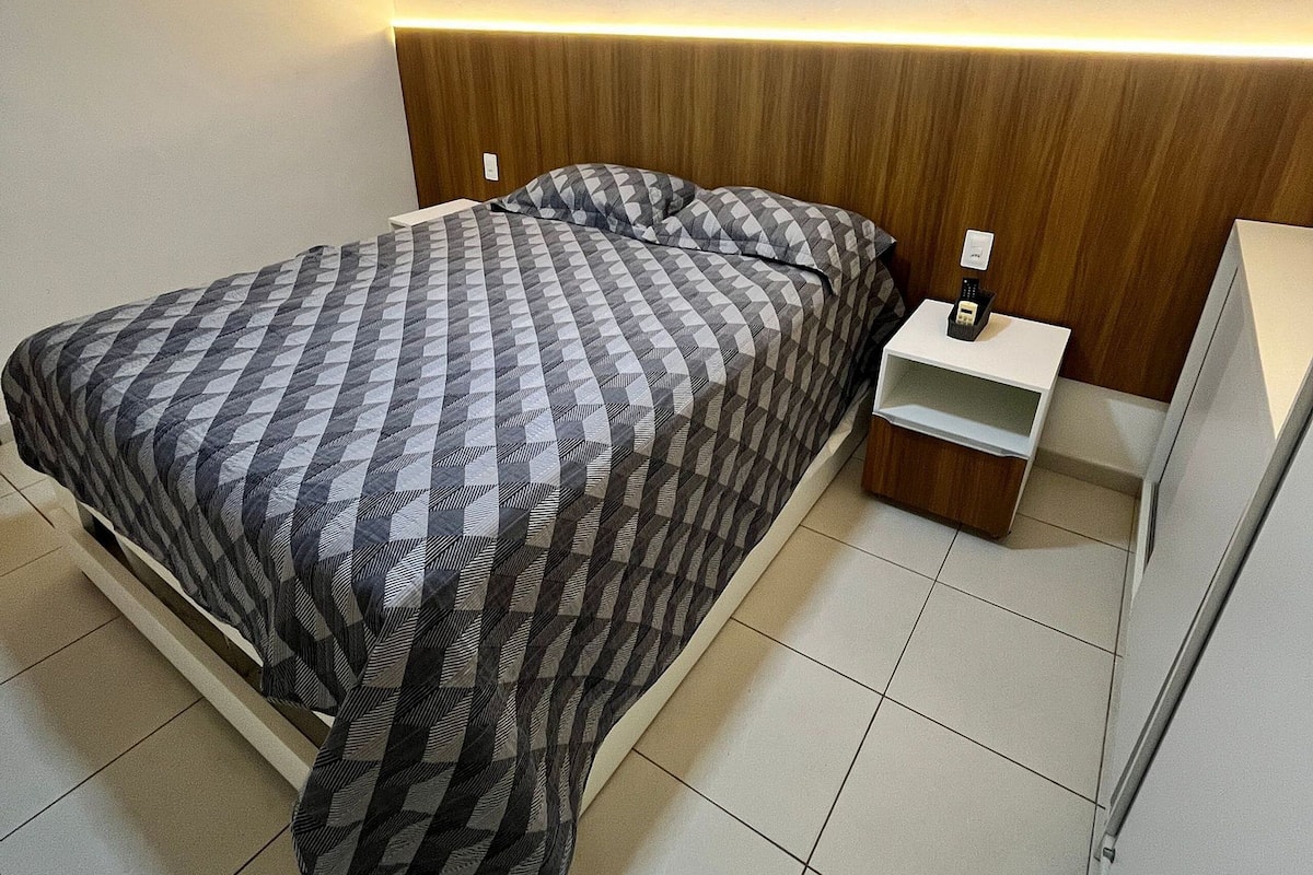 Un lit bien fait, avec une couvre-lit à carreaux, une tête de lit en bois, une table de chevet avec une lampe et une armoire blanche.