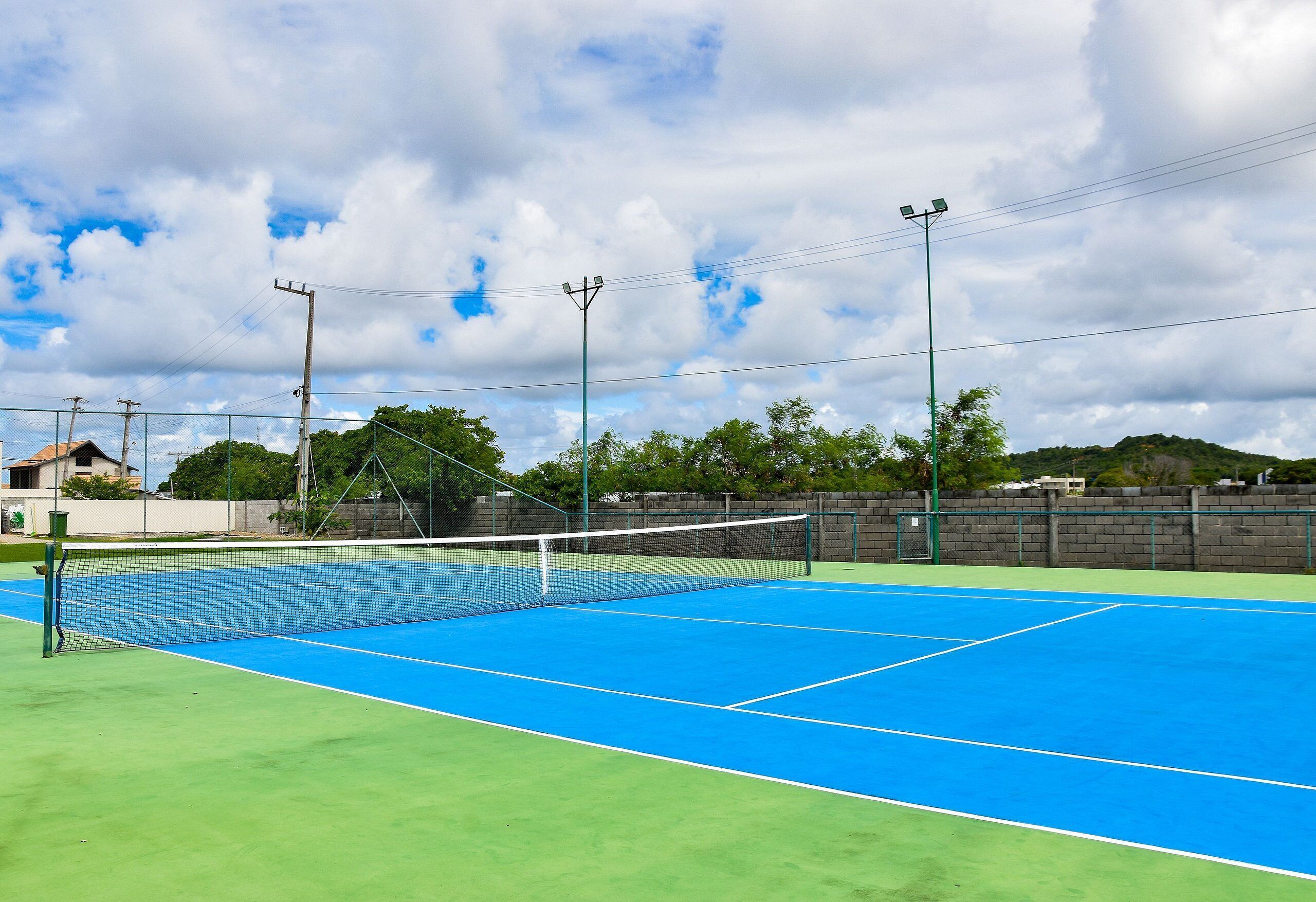 Una cancha de tenis al aire libre con una red, rodeada por una cerca verde y un muro de ladrillo.