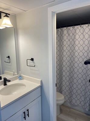 Een moderne badkamer met een wit wastafel, een zwarte kraan en een spiegel. Er is een douchegordijn met een geometrisch patroon, en een toilet is op de achtergrond zichtbaar.