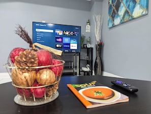 Una habitación con televisor mostrando una interfaz de servicio de transmisión, un frutero, un control remoto y un libro sobre la mesa.