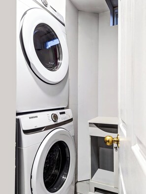 A compact laundry room with two stacked washing machines, one above the other, and a small white cabinet with a brass door. - Modern Cozy Luxury Suite in Ajax | 5 Min to Hwy 401 | Kitchen/ Laundry/Parking  (Ajax)