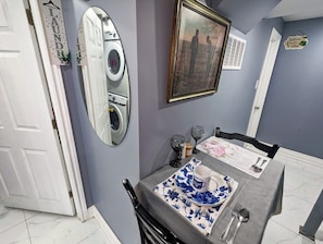 A dining area with a table set for two, a mirror reflecting a washing machine, and a framed picture on the wall.