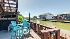 A wooden deck with chairs and a grill, overlooking a canal with buildings in the background. - Resort Perks: Waterfront Rockport Home! (Rockport)