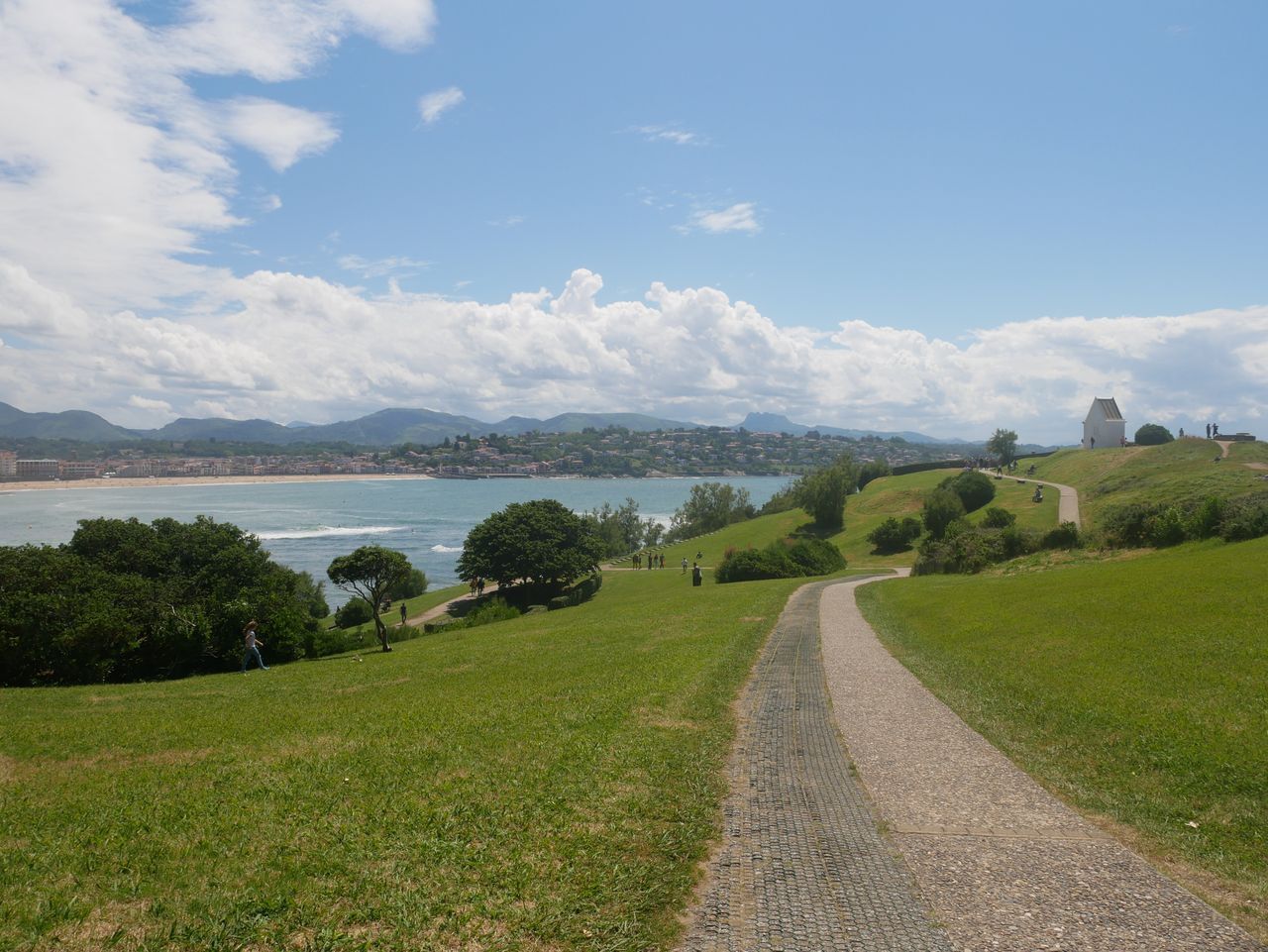 Ein gepflasterter Weg führt zu einer Küstenlandschaft mit Blick auf das Meer und entfernte Gebäude.