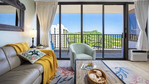 A room with a sofa, armchair, and a table with a magazine and decorative items. A large window offers a view of a green landscape and buildings. - 36FL Diamond Head & Ocean view condo w/ parking (Honolulu)