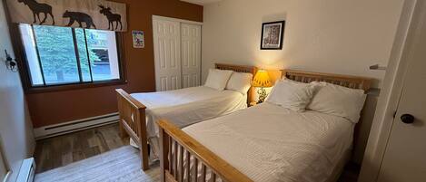 A bedroom with two beds, a window with a view of trees, a ski sign, and a framed picture on the wall.