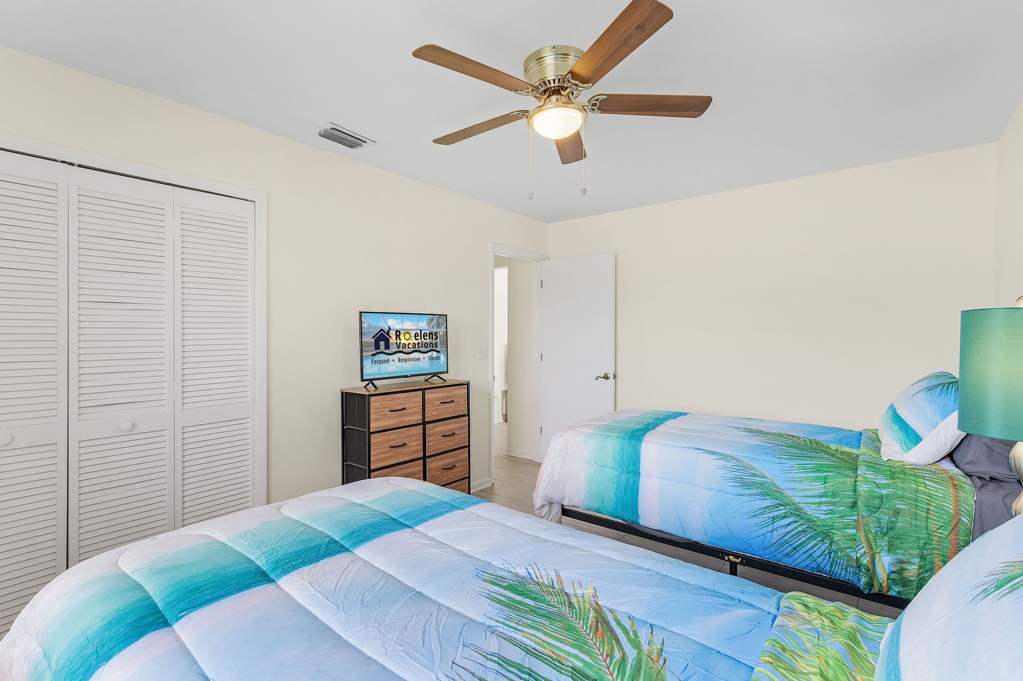 A bedroom with two beds, a ceiling fan, a wardrobe, and a sign on the dresser.