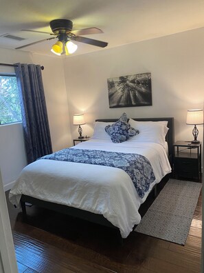 A bedroom with a bed, two bedside lamps, a ceiling fan, a window with curtains, and a painting on the wall.