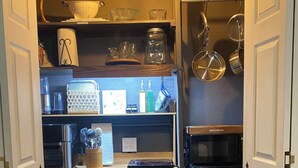 Cocina compacta con un refrigerador Galanz, microondas y cafetera. Hay estantes con distintos utensilios de cocina y una pequeña alfombra en el suelo.