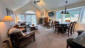 A spacious living room with a fireplace, a dining area, and large windows offering a view of the outdoors. - FV44 tastefully updated single level with AC, WiFi, minutes from skiing, Dog Friendly! (Bretton Woods)