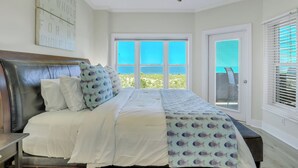 Quarto com cama, ventilador de teto, janela ampla com vista para o oceano e porta com persianas.
