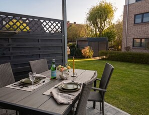 Un espace de restauration en plein air avec une table dressée pour deux personnes, entourée de chaises en osier, d’une bouteille de vin et d’une bougie.