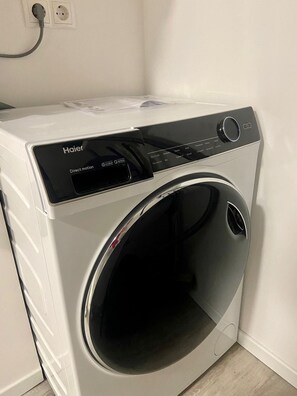 Una lavadora Haier blanca de carga frontal, con panel de control, etiqueta de movimiento directo y cable de alimentación conectado a un enchufe de pared.