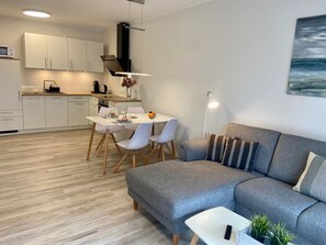 Moderni olohuone, jossa on harmaa sohva, ruokapöytä tuoleineen, taustalla keittiö ja seinällä taulu.