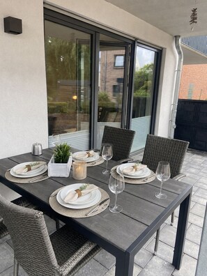 An outdoor dining area with a table set for four, surrounded by wicker chairs, and a large window providing a view of the neighborhood.