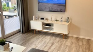 Ein modernes Wohnzimmer mit einem wandmontierten Flachbildfernseher, einem weißen Konsolentisch mit Dekoration und Blick auf einen Strand durch ein großes Fenster.