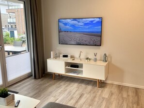 Un salon moderne avec un téléviseur à écran plat fixé au mur, une console blanche agrémentée d’objets décoratifs et une vue sur la plage, visible à travers une grande fenêtre.