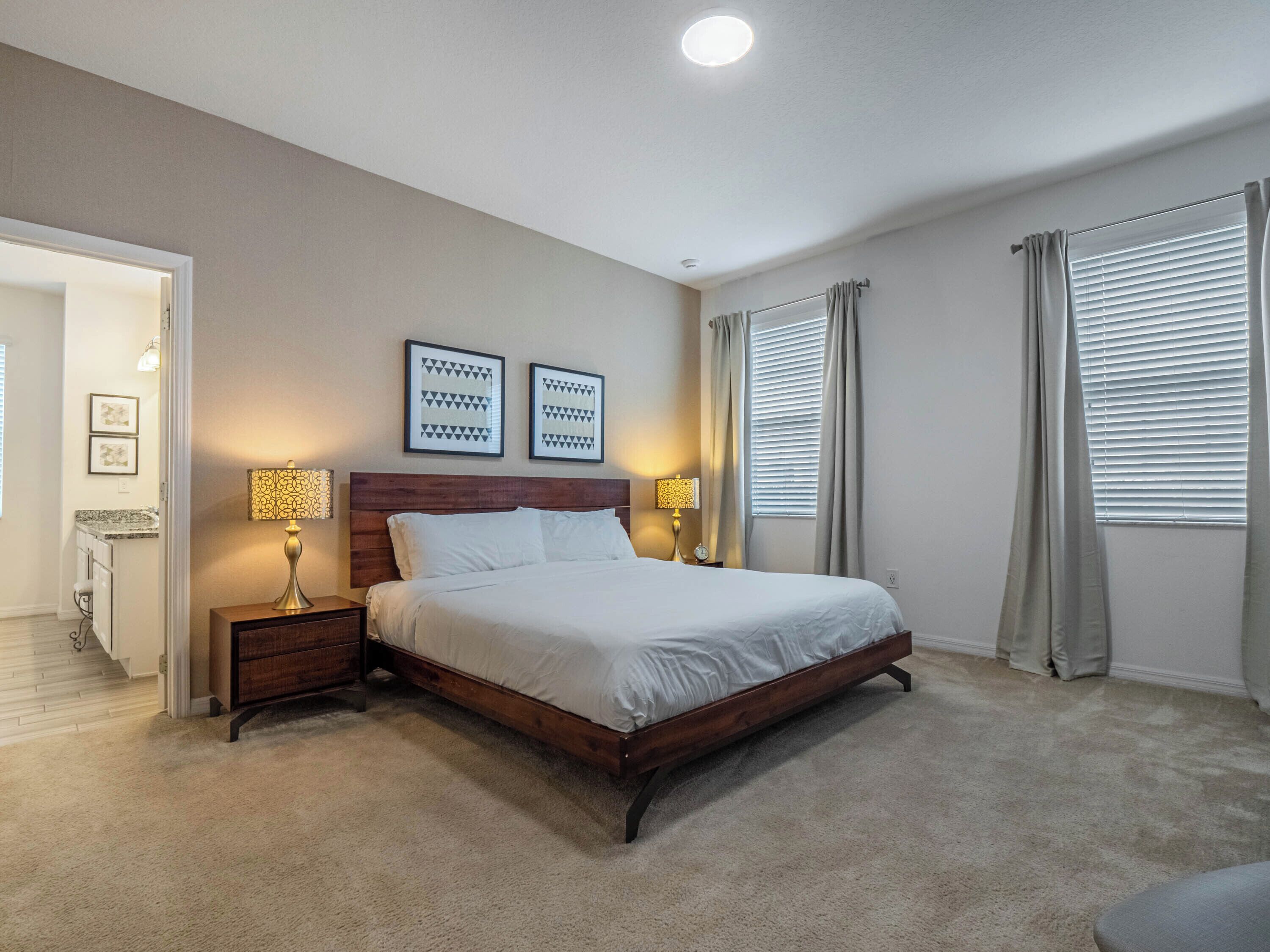 A bedroom with a wooden bed, two bedside lamps, a ceiling light, and a door leading to a bathroom.