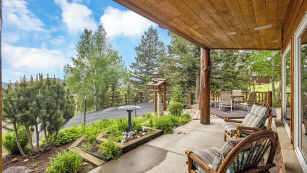 Une terrasse couverte, dotée de meubles en bois, d’une mangeoire à oiseaux et d’un jardin où poussent diverses plantes et arbres.