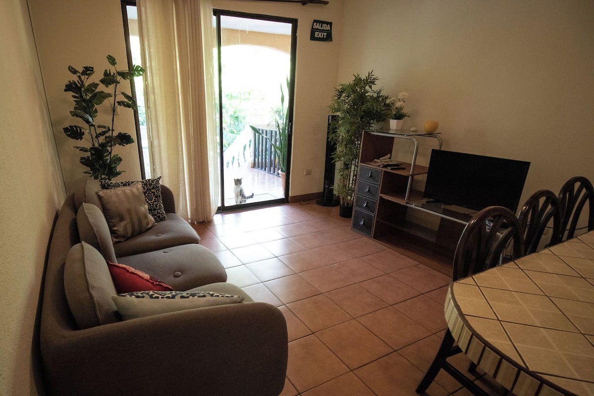 A living room with a sofa, a dining table, and a TV. There is a door leading to a balcony.