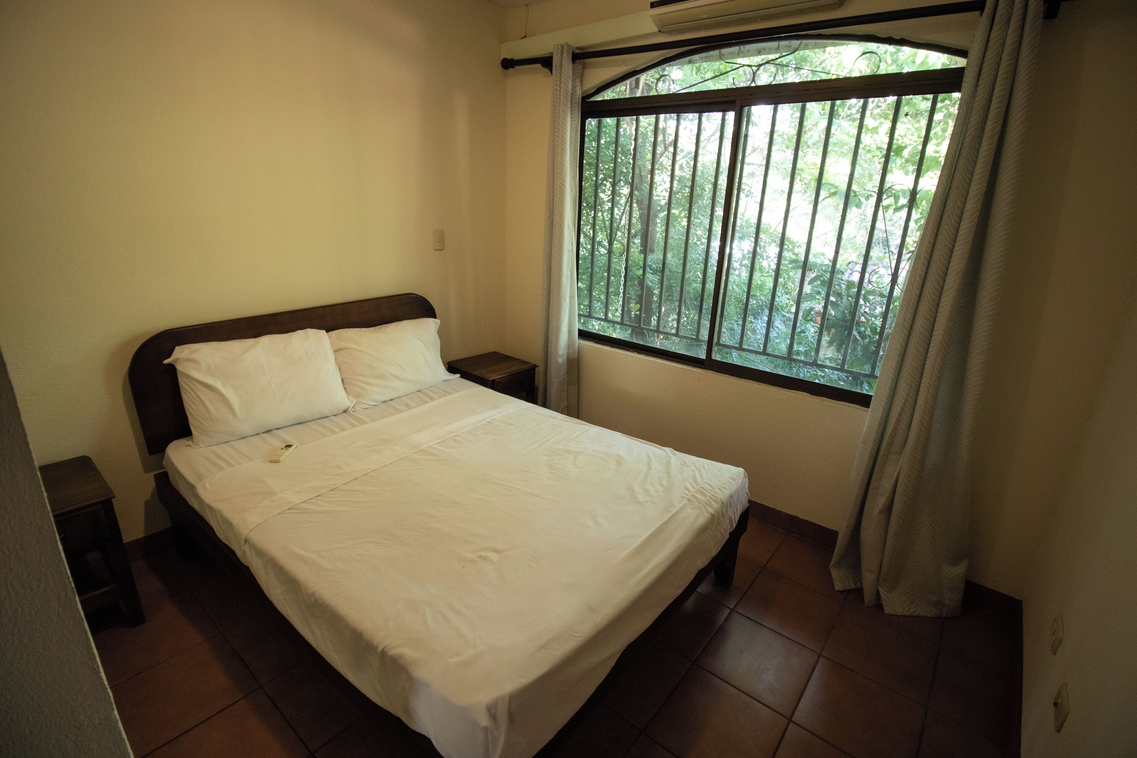 A single bed with white bedding in a room with a window and a view of greenery outside.