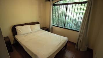 A single bed with white bedding in a room with a window and a view of greenery outside.