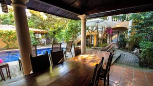 A covered outdoor dining area with a pool, chairs, and umbrellas. - Anona Guesthouse (Sardinal)
