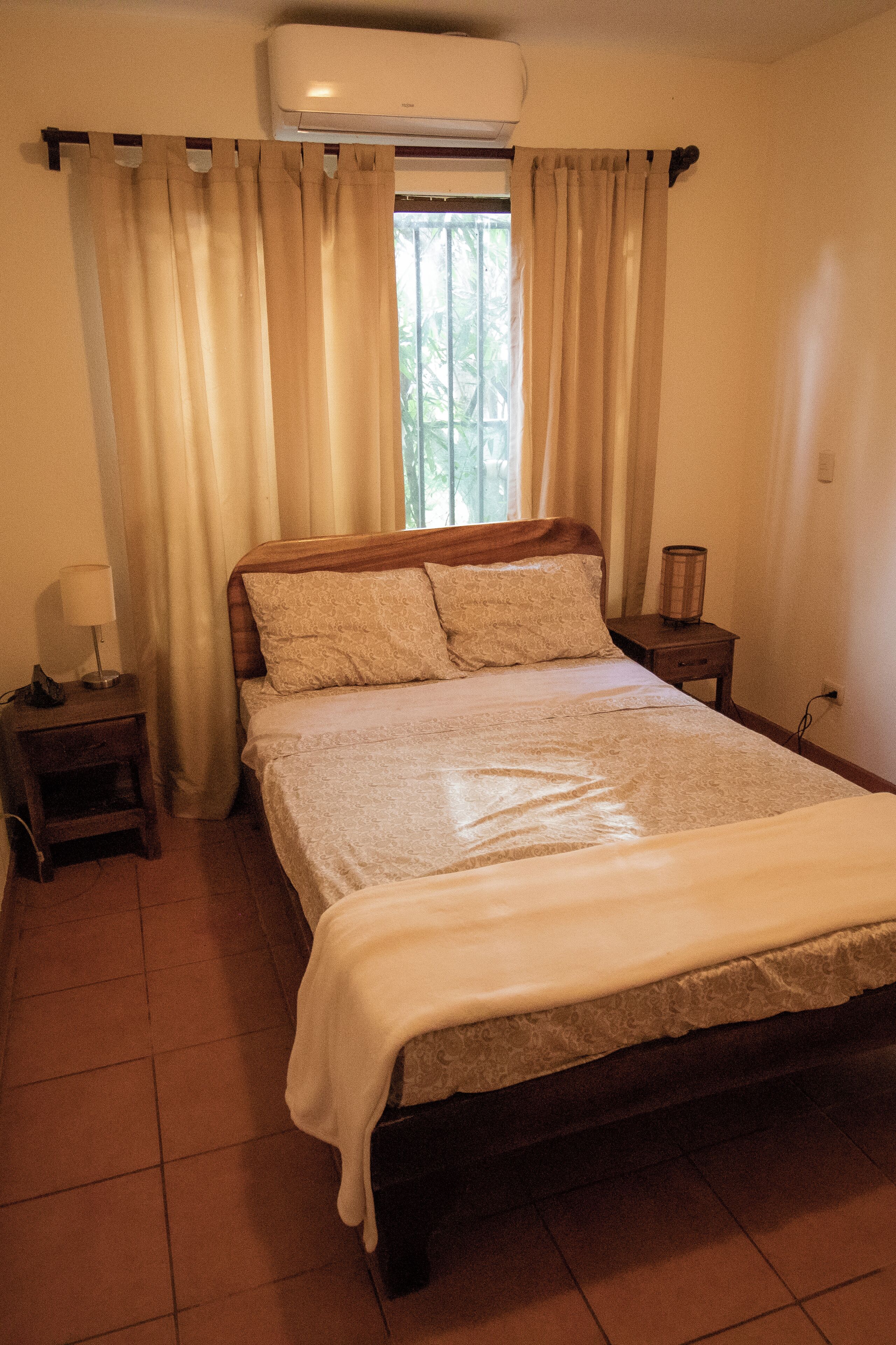 Une chambre à coucher comprenant un lit, des rideaux, une lampe de chevet, une table de chevet et un appareil de climatisation.