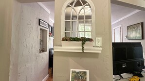 Ein Flur mit weißer Wand, Holzboden, einem gerahmten Blumenbild, einem Fenster mit weißem Rahmen, einem Willkommensschild und einem kleinen Tisch mit einem Korb.